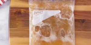 A sealed Ziploc bag filled with a brownish liquid, likely homemade broth or stock, sits on a wooden surface. There are bubbles visible within the liquid, and a patterned cloth is partially visible in the upper left corner of the image.
