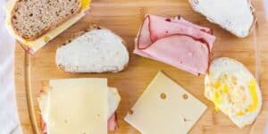 A wooden board holds ingredients for a sandwich, including two slices of bread with spread, folded slices of ham, Swiss cheese, and fried eggs. One piece of bread has cheese on top, and another has cheese and a fried egg.