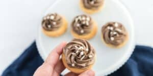 A hand holding a small cupcake topped with swirled chocolate frosting and white sprinkles. In the background, five additional similar cupcakes are arranged on a white plate, set against a soft-focus white and blue backdrop.