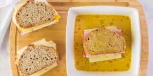 Two sandwiches on a wooden cutting board next to a shallow bowl filled with a liquid, likely an egg mixture, in which a third sandwich is partially dipped. The sandwiches consist of whole grain bread, slices of cheese, and ham.