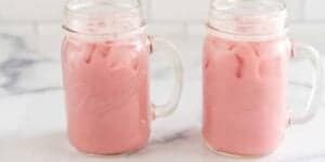 Two mason jar mugs filled with iced pink beverages are placed on a light-colored surface with a blurred background. The beverages appear creamy and cold, and the jars have handles for easy gripping.