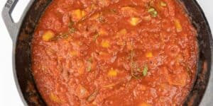 A close-up view of a cast iron skillet filled with thick, vibrant red tomato sauce, garnished with basil leaves and chunks of vegetables. The sauce appears to be simmering, displaying a rich and hearty texture.