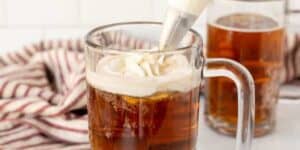 A glass mug filled with dark, fizzy liquid is topped with white foam from a piping bag. Another similar mug is in the background, and a red and white striped cloth is partially visible beside the mugs on a light-colored surface.