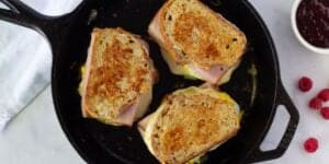 A cast iron skillet contains three grilled cheese sandwiches made with toasted bread, melted cheese, and slices of ham. To the right of the skillet, there are fresh raspberries and a small dish of raspberry jam on a white surface. A white cloth is partially visible on the left.