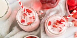 Two mason jars filled with a pink strawberry drink, garnished with strawberry slices and accompanied by red-striped straws. The jars are placed on a light fabric, with fresh strawberries and a strawberry-filled container nearby.