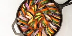 A cast-iron skillet filled with a colorful ratatouille, featuring neatly arranged slices of tomato, zucchini, eggplant, and red onion. The vegetables are placed in a circular pattern over a bed of rich tomato sauce, seasoned with pepper.