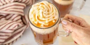 A hand holding a glass mug filled with a frothy beverage topped with a generous swirl of whipped cream and drizzled with caramel sauce. A striped cloth and another similar mug are visible in the background.