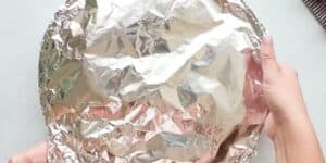 A person holding a circular baking dish covered with crinkled aluminum foil with both hands, positioned centrally in the image against a neutral background. The foil cover creates a shiny and reflective surface, showing slight dents and folds.