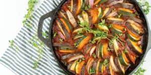 A cast-iron skillet filled with beautifully arranged slices of zucchini, eggplant, and tomato in a vibrant red sauce, garnished with fresh thyme sprigs. The skillet rests on a white and blue striped cloth.