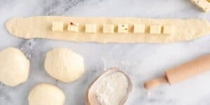 A marble countertop with dough balls, a rolling pin, and a wooden scoop filled with flour. A rolled-out dough strip with evenly spaced cheese cubes is placed horizontally across the middle, ready for preparation. This Jalapeño Cheese Stuffed Pretzel Recipe Inspired by Disney promises a delightful treat.