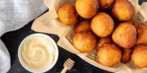 A basket filled with golden brown fried balls of dough, inspired by a famous Disney restaurant's fried biscuits recipe, sits on parchment paper. Beside it, a small white bowl holds creamy sauce, and a wooden honey dipper rests nearby. A black and white checkered cloth is partially visible on the side.