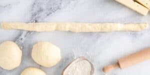 A partially rolled-out dough stick is in the center of a marble countertop, surrounded by three dough balls, a small wooden bowl of flour, and a wooden rolling pin. Inspired by Disney's Jalapeño Cheese Stuffed Pretzel recipe, there's also a stack of beige blocks in the top right corner.