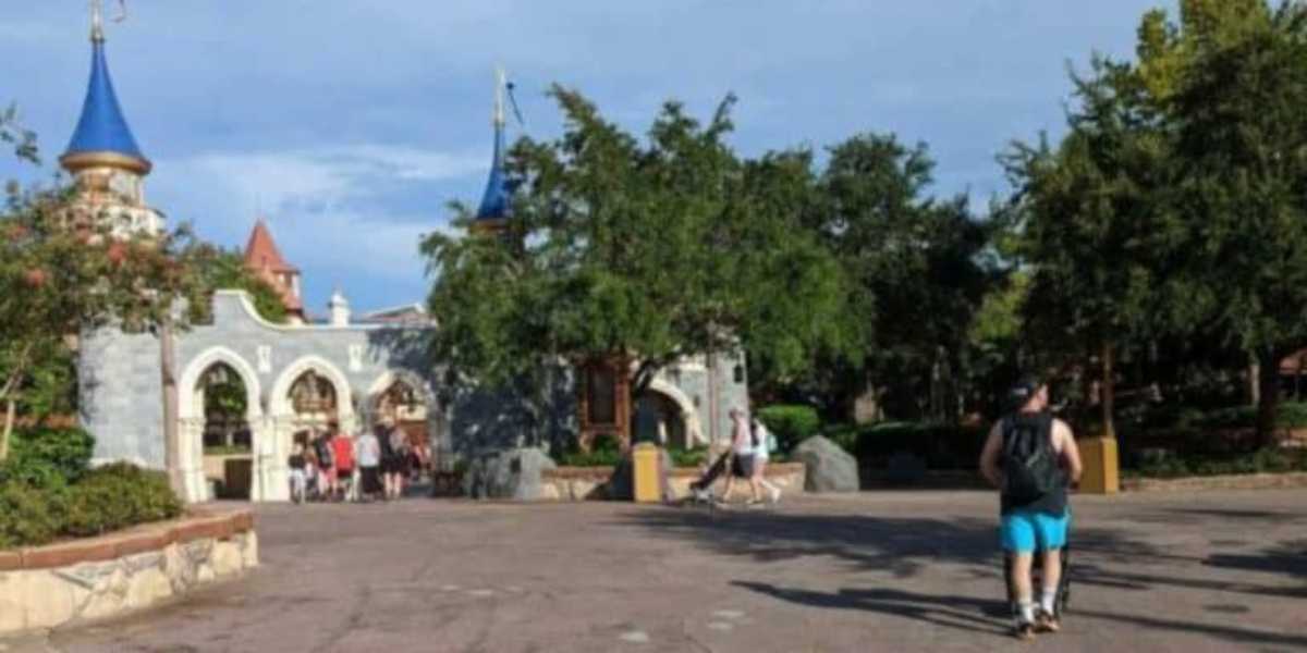 Park visitors walking on a paved path towards a castle-like structure with blue conical roofs and a stone facade. Trees and greenery are on either side, with clear, blue sky above. A person with a backpack and shorts is prominently walking in the foreground.