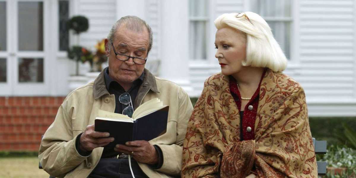 An elderly man with glasses reads a book while an elderly woman with blonde hair and a patterned shawl looks at him attentively. They are seated on a bench in front of a white house with red brick steps. Both appear to be engaged in a calm, shared moment.