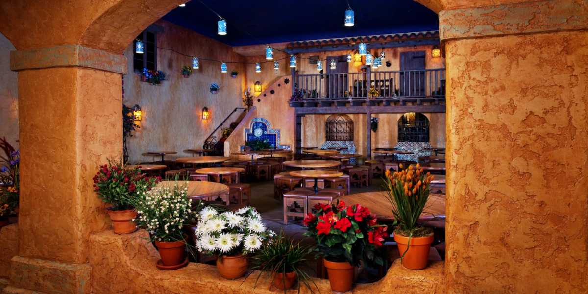 A cozy, rustic indoor courtyard at Pecos Bill Tall Tale Inn and Cafe features terracotta walls and wooden beams. Potted flowers line the foreground while round wooden tables with benches fill the open space. Hanging lanterns cast a warm glow, and a staircase leads to a wooden balcony adorned with more flowers.