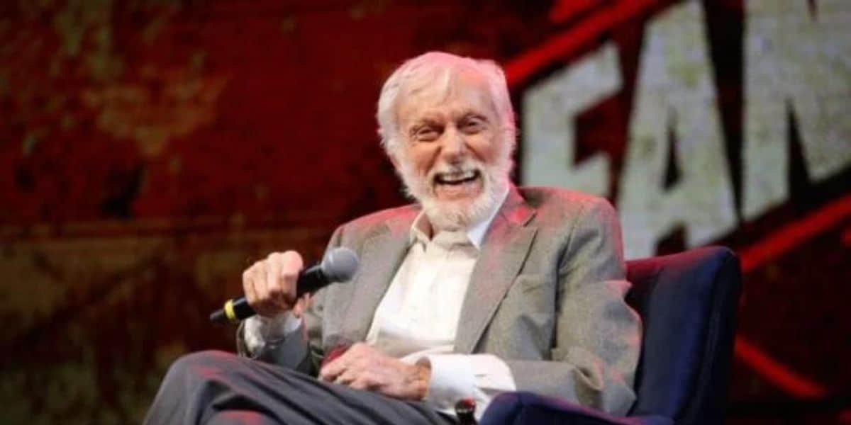 An elderly man with white hair and a beard, wearing a grey suit and light-colored shirt, sits on a blue chair while holding a microphone and laughing. The background appears to have a graphic design with bold text partially visible.