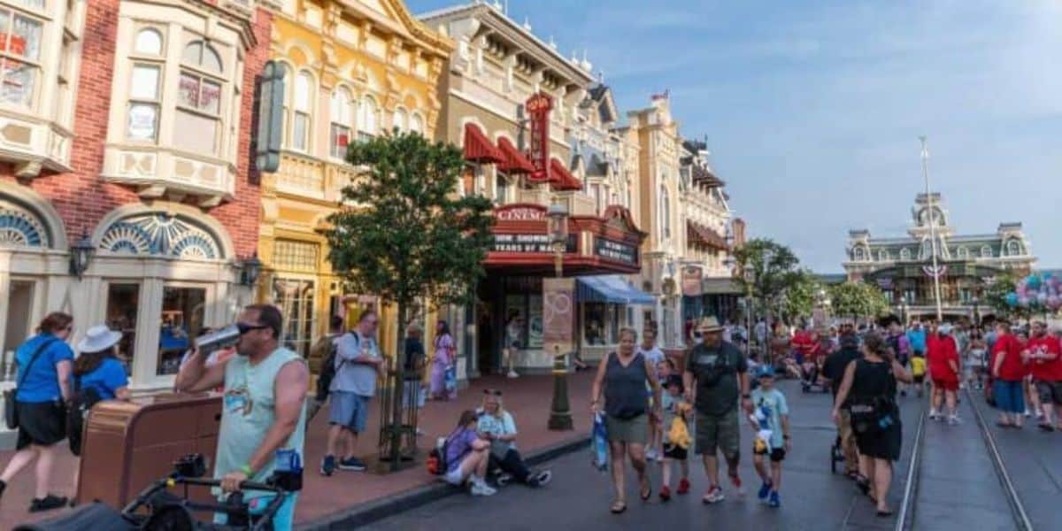 A busy street in a theme park with people walking, some holding balloons or pushing strollers. Colorful buildings line the street, including an old-fashioned theater marquee. In the distance, there's a large building with a clock and flag on top.
