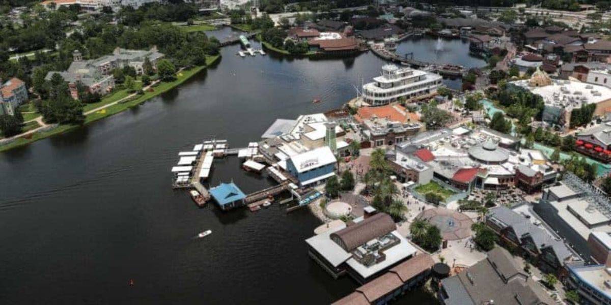 Aerial view of a lively, waterfront shopping and dining district. Various buildings with different roof styles are clustered around the water. Boats are docked along the waterfront, and green spaces with trees are visible in the background.