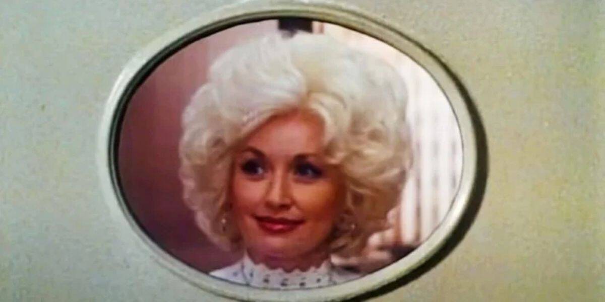A person with platinum blonde, curly hair looks through a small, rounded window with a light-colored frame. The individual is wearing a white, lacy high-collared blouse and has a pleasant expression on their face. The partial background features soft, warm lighting.