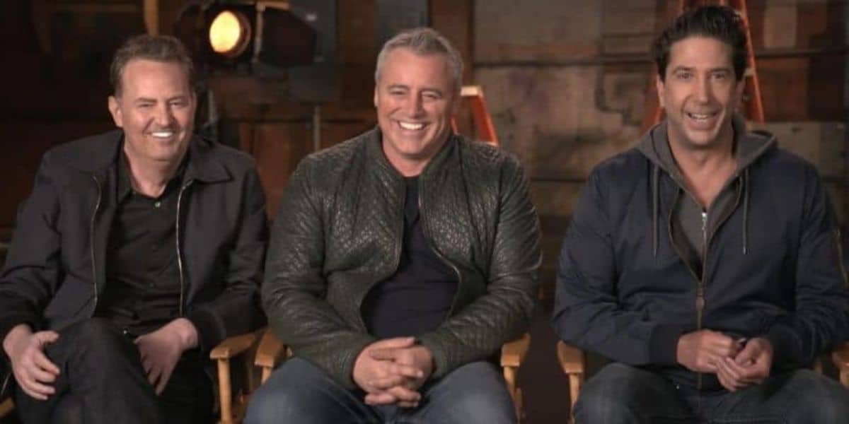 Three men are sitting on director's chairs, smiling. They are in a studio setting with dark, blurred background elements like lights and equipment. They are casually dressed in jackets and shirts.