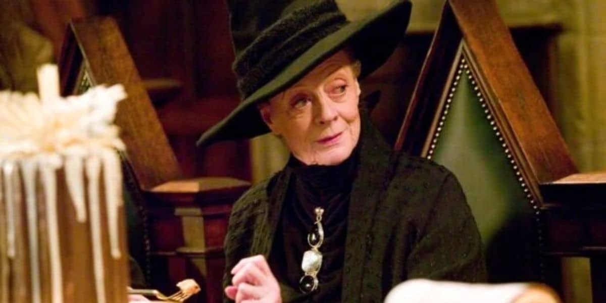An elderly woman with a serious expression sits at a desk, dressed in a black robe and a tall, wide-brimmed black hat. She has a long necklace with a large pendant. The background features wooden furniture and a large candle.