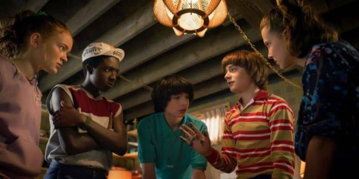 A group of five children stand in a room lit by a ceiling lamp. They are engaged in a conversation. Two of the children are wearing striped shirts, one is wearing a teal shirt, while another is in a tank top and a sailor hat. The room has a wooden ceiling.