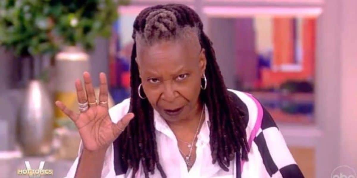 A person wearing a white shirt with a patterned sleeve is seen raising their hand slightly while looking into the camera. They have long dreadlocks and are in an indoor setting with blurred background elements.
