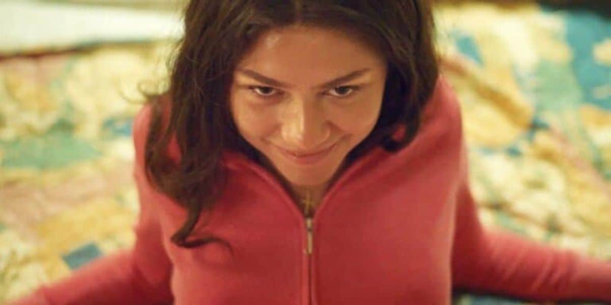 A person with long, dark hair is sitting on a colorful, patterned surface. They are wearing a pink zip-up top and looking up at the camera with a subtle smile.