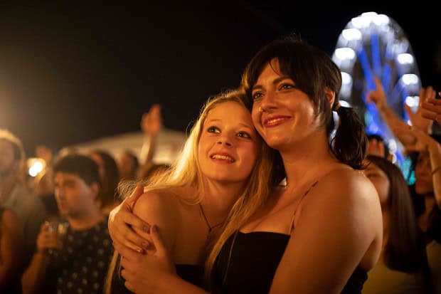Amidst the lively and festive night event, two people embrace and smile in the crowd. A ferris wheel lights up the scene like a Disney Plus film backdrop. The atmosphere is enchanting, capturing a moment that feels worlds away from Avetrana or Hollywood glamour.