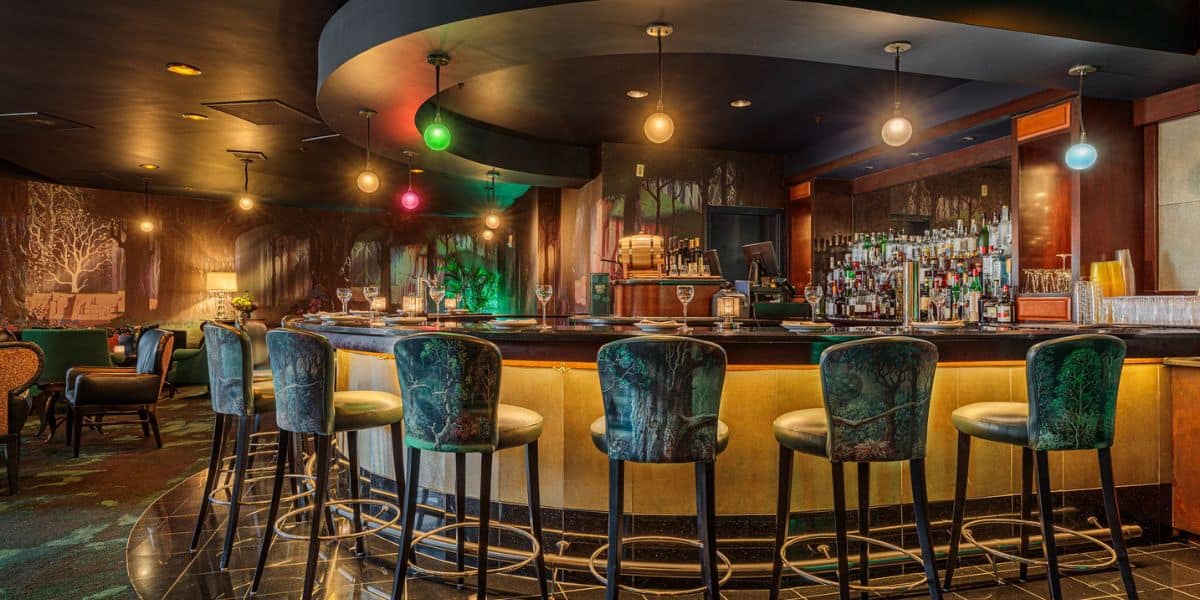 A dimly lit bar with green cushioned barstools around a curved counter. The backdrop features trees and a stocked bar with bottles. Hanging spherical lights add to the cozy atmosphere, with a few tables and chairs visible in the background.