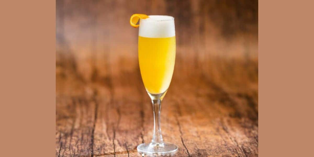 A tall champagne flute filled with a yellow-orange cocktail, garnished with a twist of orange peel on the rim, set against a warm, wooden background.