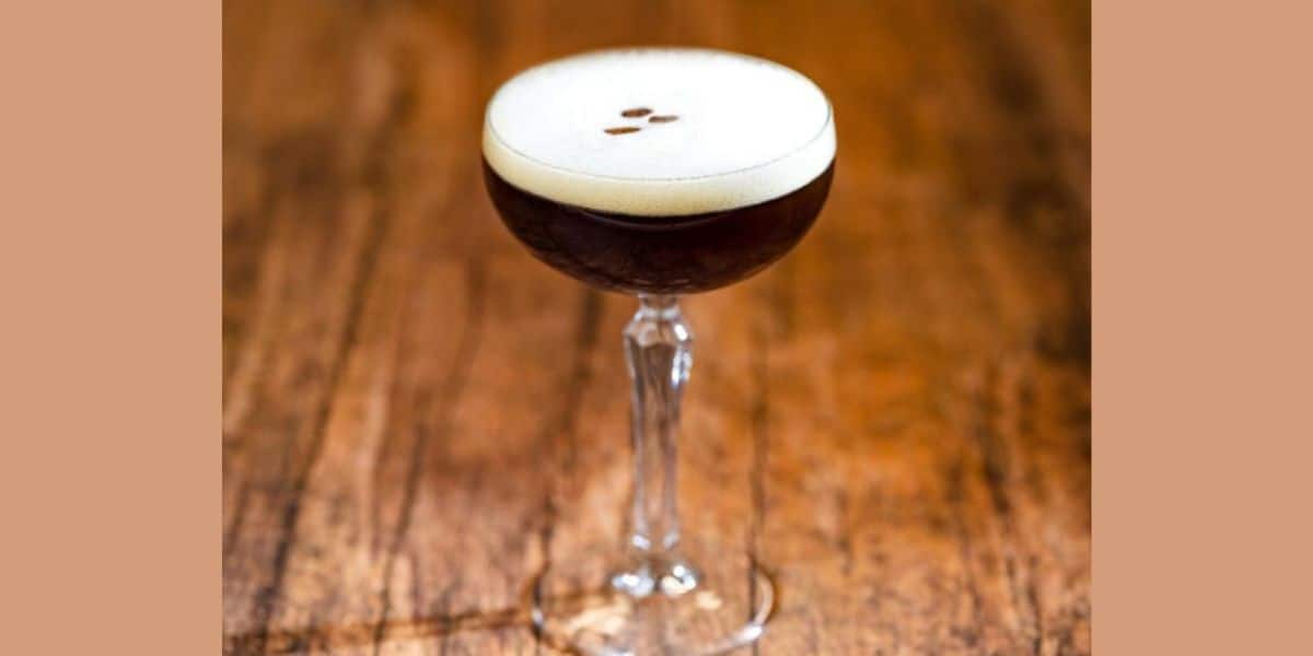 A cocktail glass filled with a dark espresso martini, topped with a creamy foam and three coffee beans, sits on a rustic wooden surface.