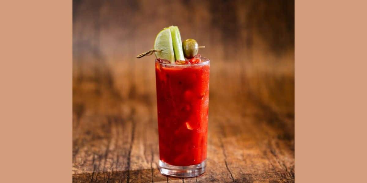 A tall glass of a red cocktail garnished with lime slices and an olive on a skewer, set against a rustic wooden background.