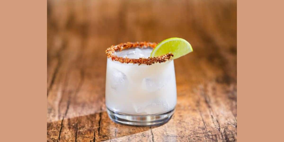 A glass filled with a light-colored cocktail is garnished with a lime wedge. The rim is coated with a red spice mix, and the drink contains ice cubes. The glass is placed on a wooden surface.
