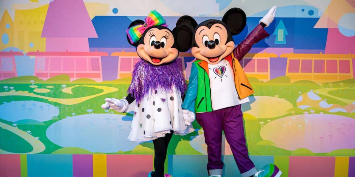 Mickey and Minnie Mouse stand together in colorful outfits, featuring rainbow accents. Minnie wears a large rainbow bow and a sequined dress. They smile against a vibrant backdrop of abstract shapes and colors.