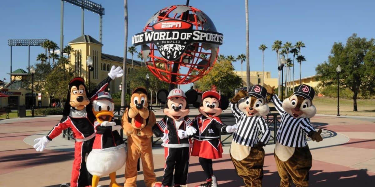 Mickey Mouse and friends, including Goofy, Donald Duck, and Chip and Dale, in sports attire, pose in front of the ESPN Wide World of Sports Complex globe. Sunny day with clear skies and trees in the background.