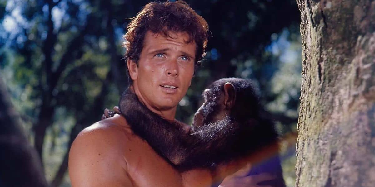 A shirtless man stands outdoors, holding and looking at a chimpanzee that clings to him. They are surrounded by trees, with sunlight filtering through the foliage. The man appears concerned or focused.
