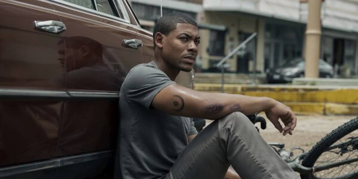 A man sits on the ground leaning against a car, with a serious expression. He wears a gray T-shirt, beige pants, and has a tattoo on his arm. A bicycle lies beside him, and the background shows an urban street scene.
