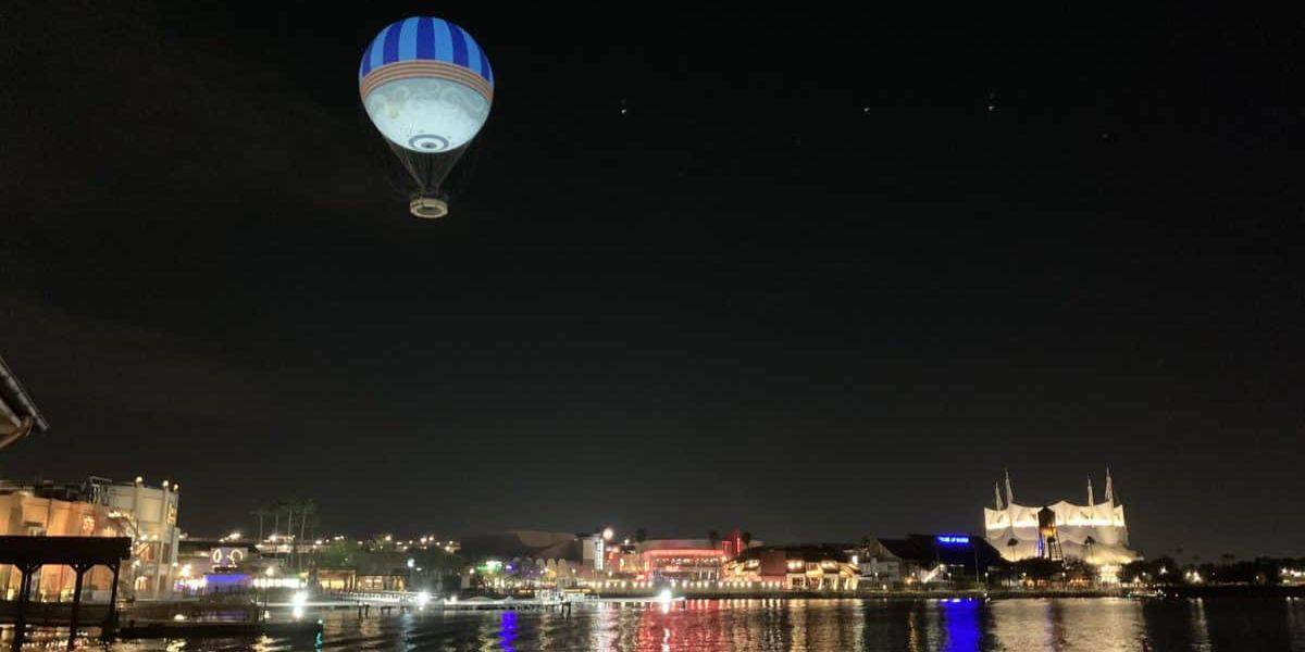 A glowing hot air balloon hovers over a cityscape at night, with buildings reflecting off a calm body of water. The scene is illuminated by various colorful lights, creating a serene and picturesque atmosphere.