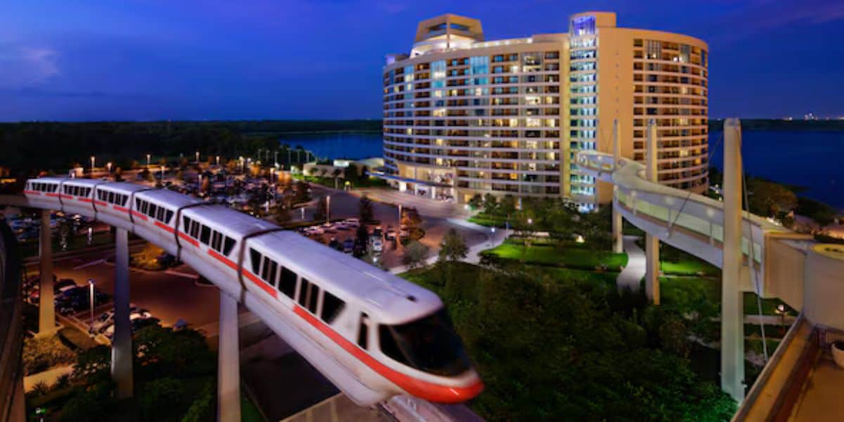 A modern monorail with a red stripe glides along a track in front of a curved, high-rise hotel lit up against a twilight sky. The hotel overlooks a large body of water, and greenery surrounds the building.