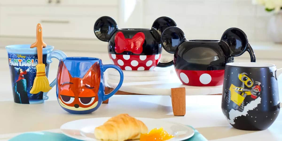 An assortment of Disney-themed mugs on a table, featuring designs from Mickey Mouse, Minnie Mouse, Zootopia, and WALL-E. A croissant and orange jam are in the foreground.
