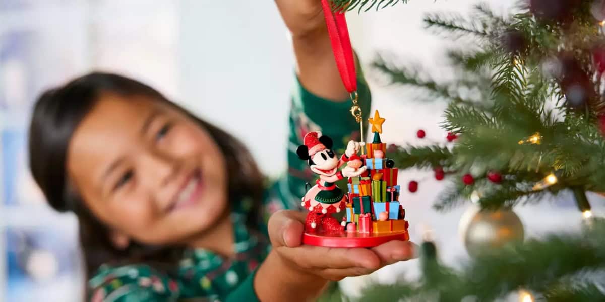 A young girl, smiling in the background, is holding a Christmas ornament depicting a character dressed in holiday attire next to a small decorated Christmas tree. The tree in the foreground is adorned with baubles.