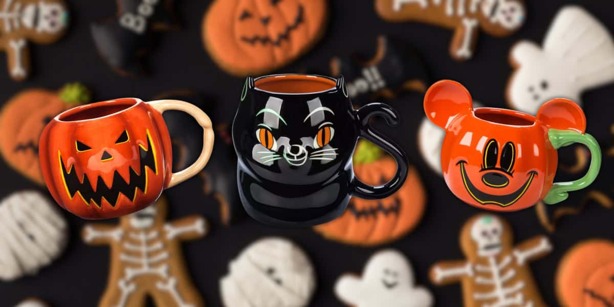 Three Halloween-themed mugs: a pumpkin face, a black cat, and a Mickey Mouse pumpkin. They are arranged in front of a blurred background featuring Halloween cookies shaped like ghosts and skeletons.