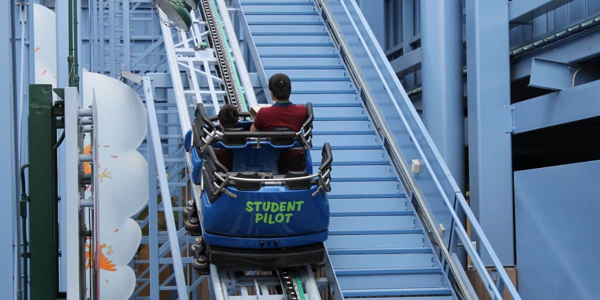 Two people in a blue roller coaster car labeled "Student Pilot" ascending a track inside a blue structure on Goofy's Sky School. The tracks and stairs are visible around them.