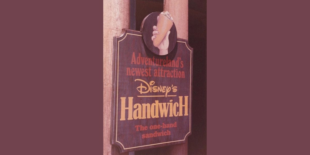 A vintage sign for Disney's Handwich at Adventureland, featuring an image of a hand holding a sandwich. The sign promotes it as "Adventureland's newest attraction" and describes it as "The one-hand sandwich.