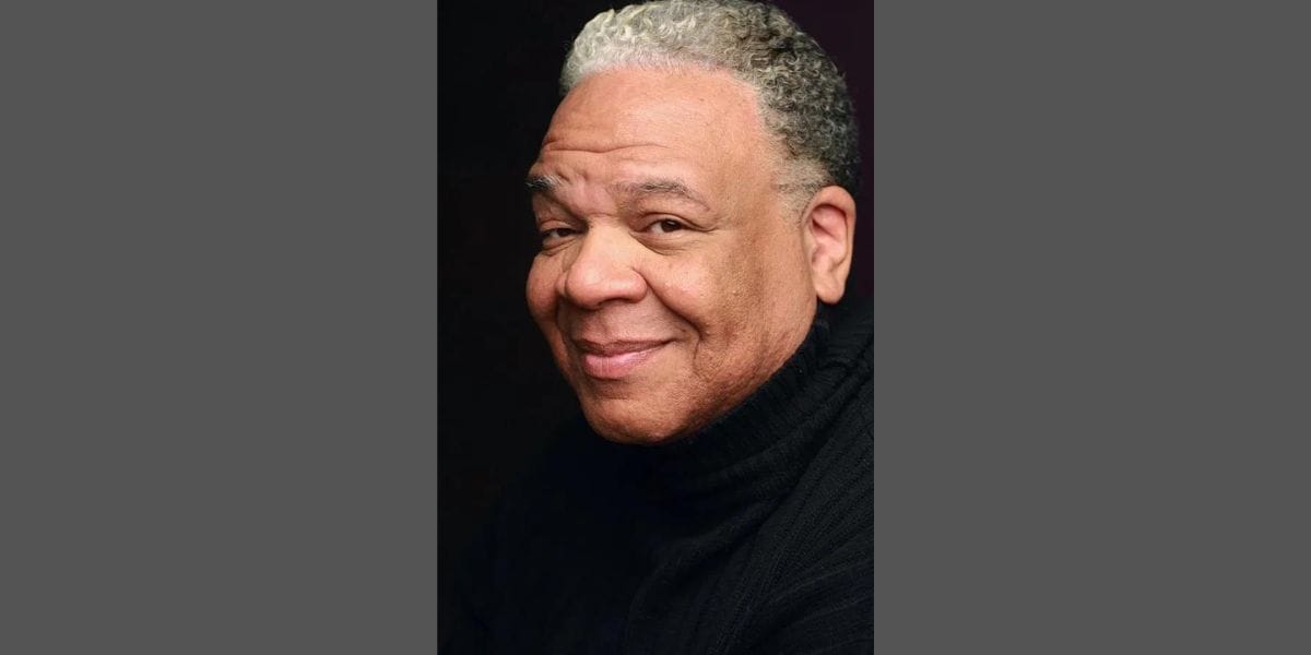 A middle-aged man with short curly gray hair and brown skin is smiling warmly. He is wearing a black turtleneck sweater and is set against a dark background. The lighting highlights his face, giving a soft, friendly appearance.
