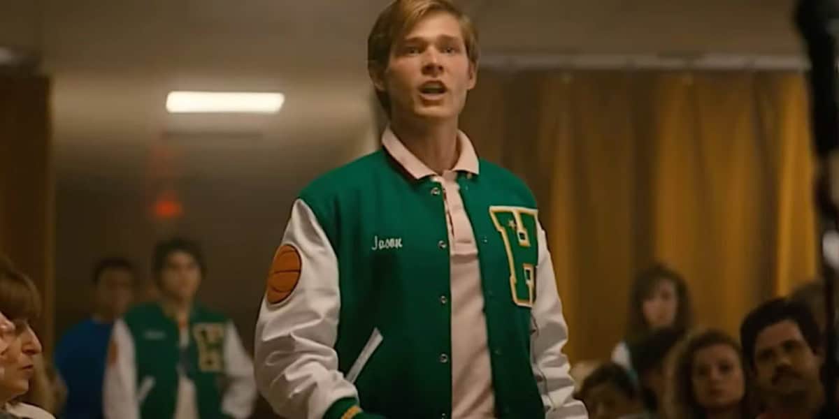 A young man in a green and white varsity jacket stands speaking passionately in a room with people sitting and watching him. The jacket has a basketball patch on the left sleeve and the name "Jamie" embroidered on it. The scene appears indoors.
