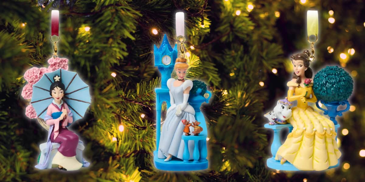 Three Disney princess ornaments hang on a Christmas tree. Mulan holds a parasol, Cinderella has a castle backdrop, and Belle is with a teacup and Chip. They are surrounded by green branches and small twinkling lights.