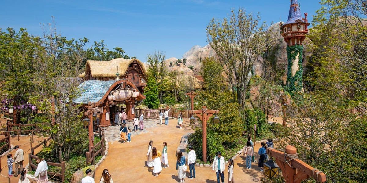 A whimsical theme park scene featuring a fairy-tale cottage and Rapunzel's tower amidst lush greenery. Visitors stroll along a winding path, enjoying the sunny day and the enchanting surroundings.