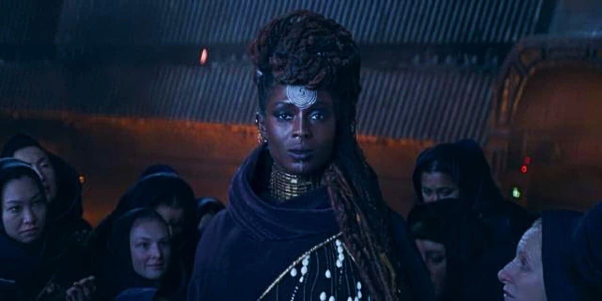 A person with intricate braided hair and a silver forehead accessory stands in the center, surrounded by a group of people wearing dark hooded garments. The background is dimly lit, conveying a mysterious and atmospheric setting.